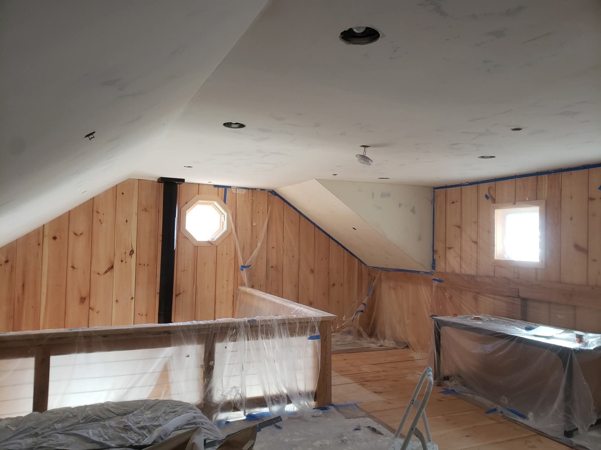 Attic loft under renovation with wood walls, sloped white ceilings, and plastic-covered railings and furniture.